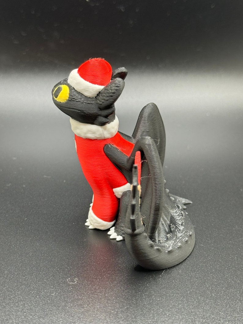 May include: A black and red 3D printed figurine of a dragon wearing a Santa hat and a red and white suit. The dragon is looking up and has a yellow eye.