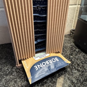 Może przedstawiać: Jasnobrązowy, pionowy pojemnik na kapsułki do kawy z rowkowaną konstrukcją. Pojemnik dozuje pojedyncze kapsułki do kawy, z widoczną etykietą "CAFFE BORBONE". Kapsułki są niebiesko-białe.