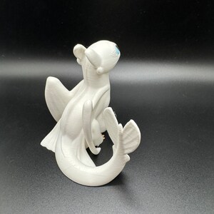 May include: White figurine of a dragon with blue eyes, wings, and a curled tail. The dragon is posed in a sitting position with its head tilted up.