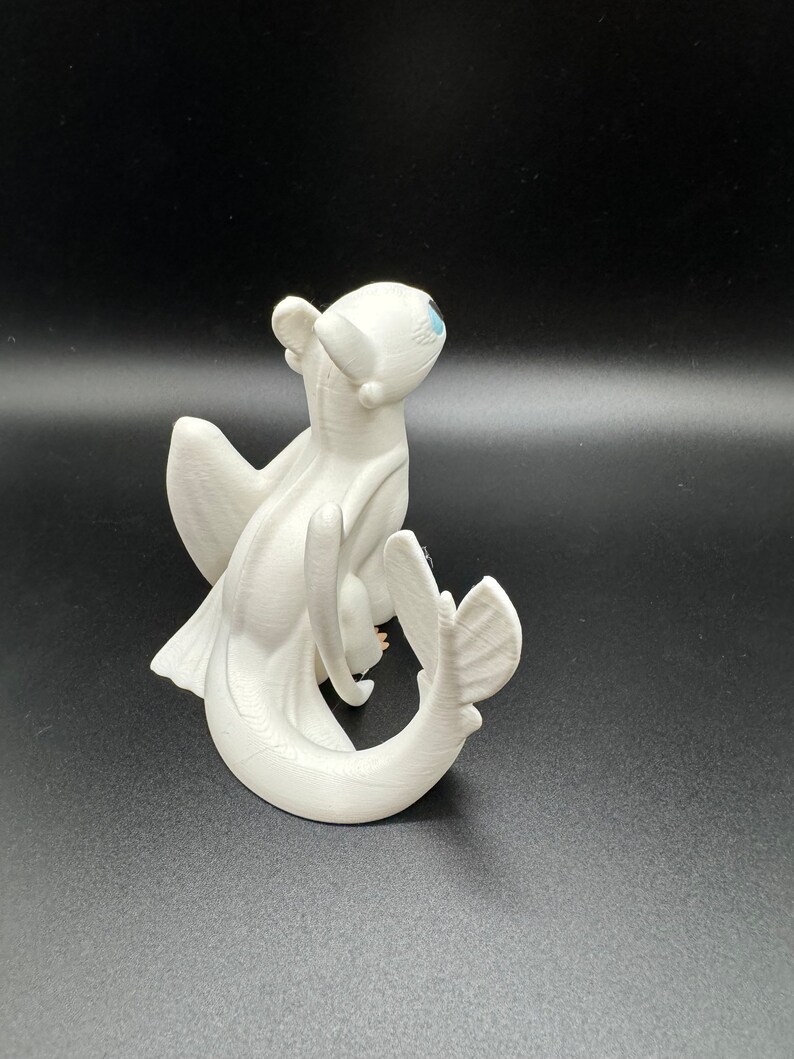 May include: White figurine of a dragon with blue eyes, wings, and a curled tail. The dragon is posed in a sitting position with its head tilted up.