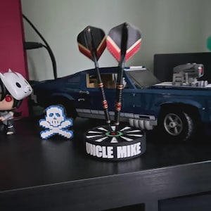 May include: A dart holder with two darts, featuring black, red, and yellow flights. The holder is black with the text "UNCLE MIKE" and a target design. A pixelated skull and a blue toy car are also visible.