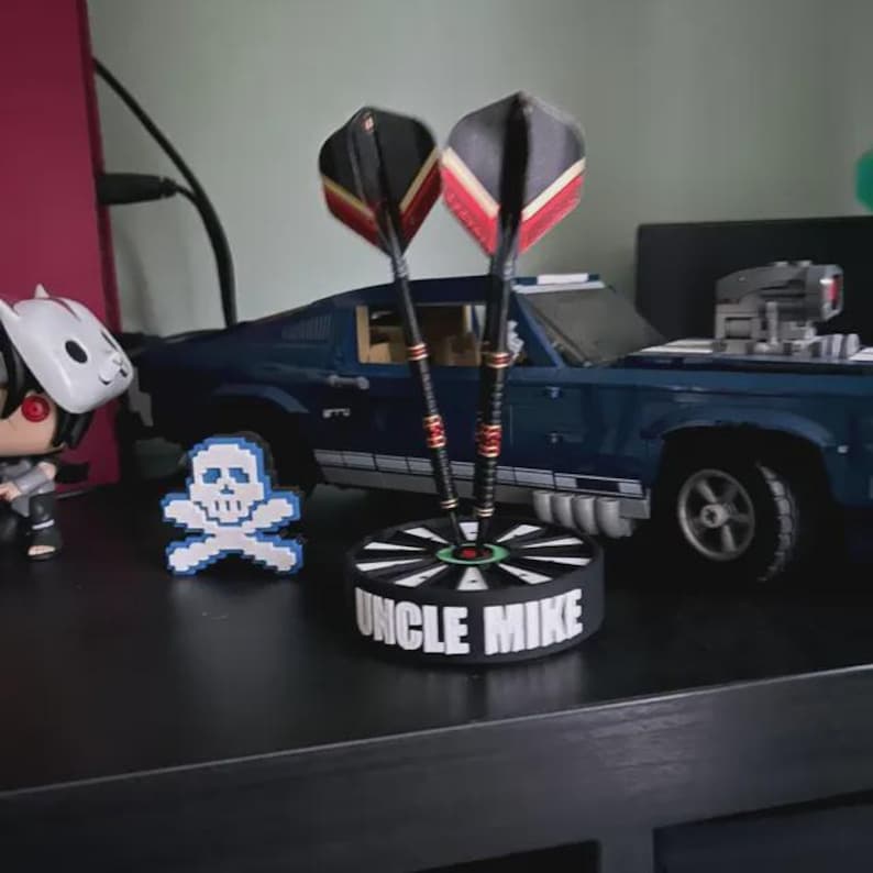 May include: A dart holder with two darts, featuring black, red, and yellow flights. The holder is black with the text "UNCLE MIKE" and a target design. A pixelated skull and a blue toy car are also visible.