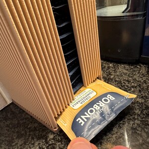 May include: A tan cardboard coffee pod holder with multiple slots, holding individual coffee pods. A hand is pulling out a coffee pod with the text "BORBONE CAFFE" and "CIALDA COMPOSTABILE" printed on it.
