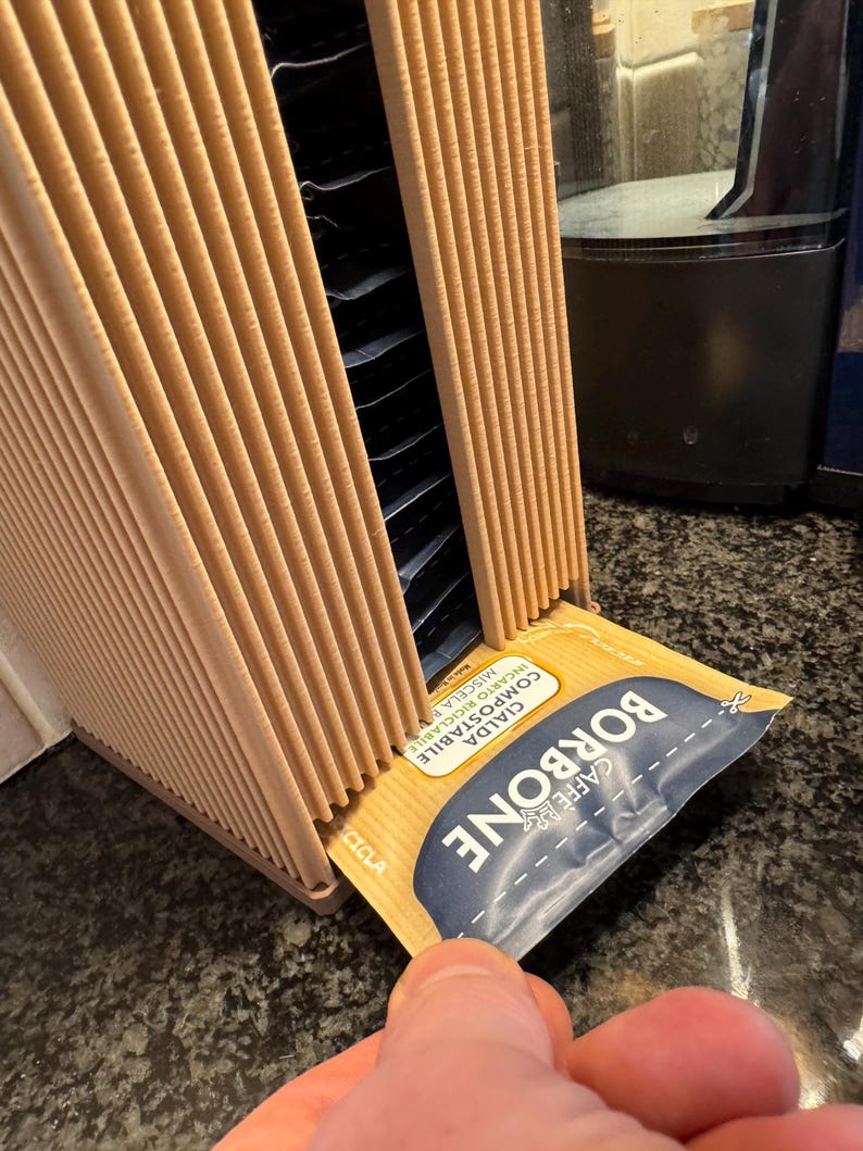 May include: A tan cardboard coffee pod holder with multiple slots, holding individual coffee pods. A hand is pulling out a coffee pod with the text "BORBONE CAFFE" and "CIALDA COMPOSTABILE" printed on it.