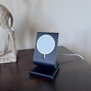 May include: A black and silver wireless charging stand with a white charging pad. The stand is on a dark brown wooden surface. A white cable extends from the stand. The stand is designed to hold a phone.