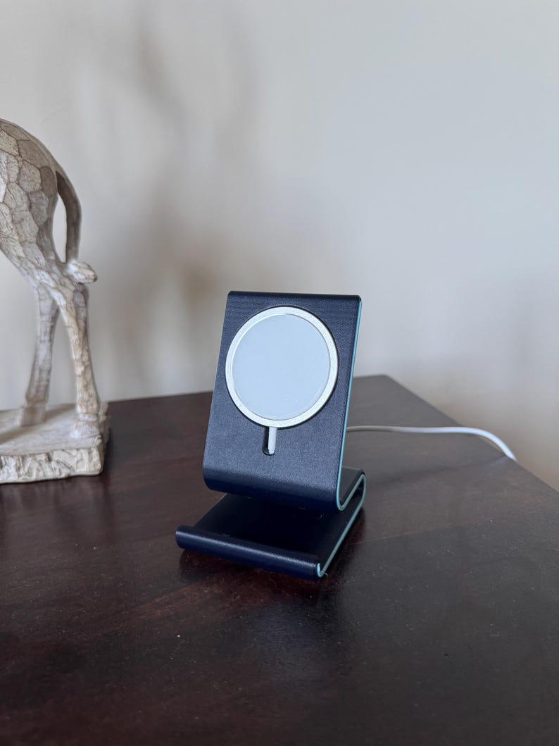 May include: A black and silver wireless charging stand with a white charging pad. The stand is on a dark brown wooden surface. A white cable extends from the stand. The stand is designed to hold a phone.