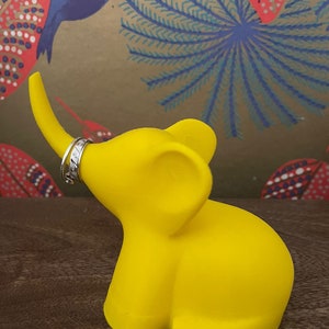 Op de afbeelding: Een felgele olifant ringhouder met een zilveren ring in zijn slurf. De olifant staat op een houten oppervlak.