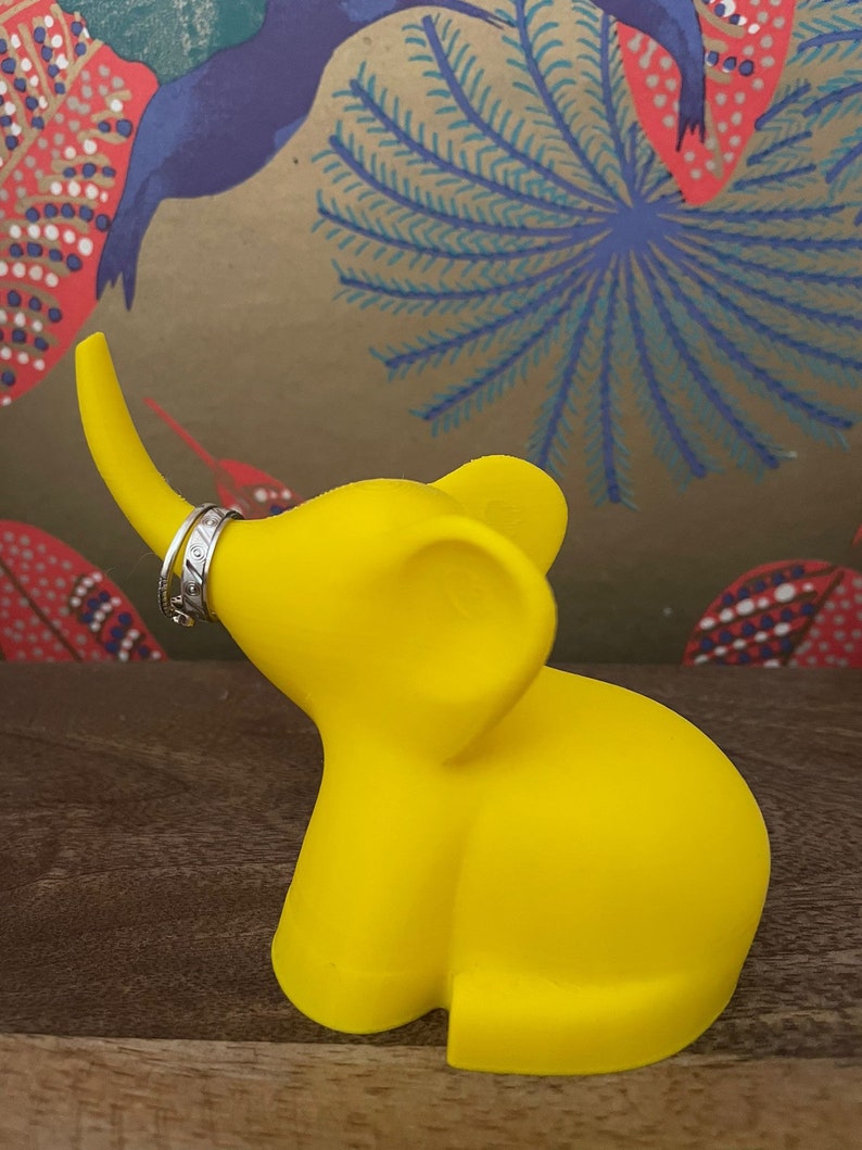 Op de afbeelding: Een felgele olifant ringhouder met een zilveren ring in zijn slurf. De olifant staat op een houten oppervlak.