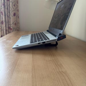 May include: A silver laptop with a black keyboard is open on a wooden desk. The screen displays a dark image. The laptop is slightly elevated by a black stand. A desk lamp and curtain are visible in the background.