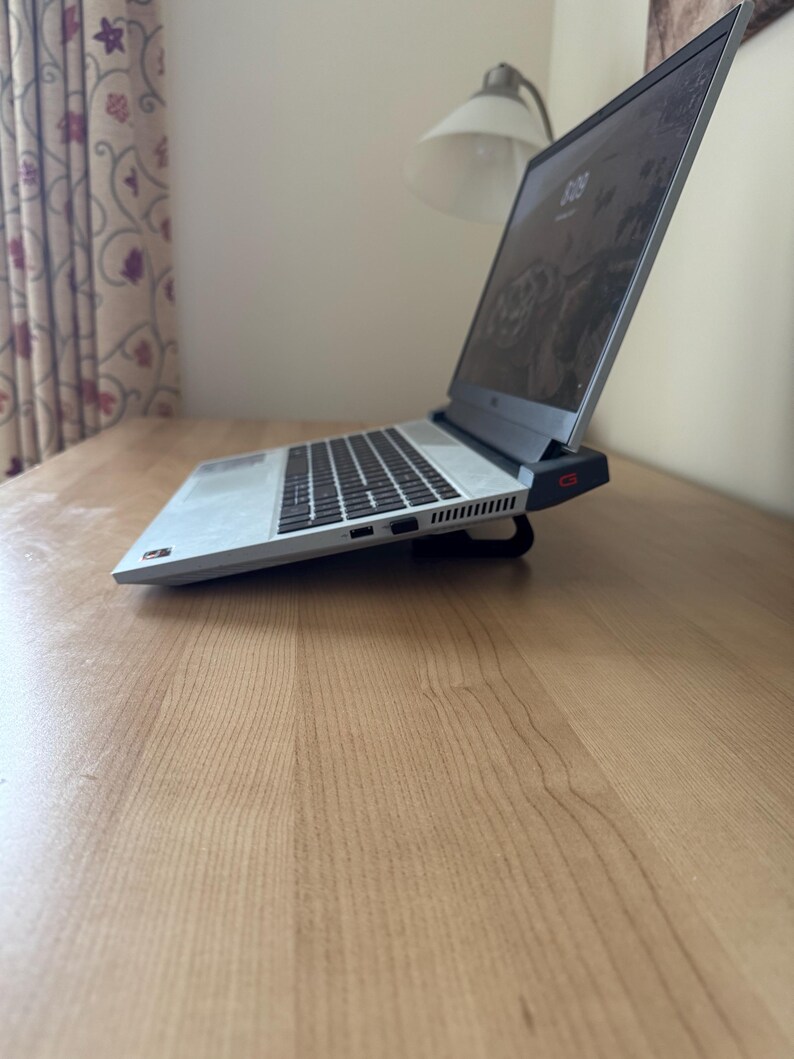 May include: A silver laptop with a black keyboard is open on a wooden desk. The screen displays a dark image. The laptop is slightly elevated by a black stand. A desk lamp and curtain are visible in the background.