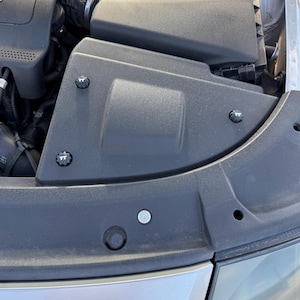 May include: A close-up view of a car engine compartment. The image shows a black plastic air intake box, silver metal components, and a red bar. The engine components are secured with silver bolts.