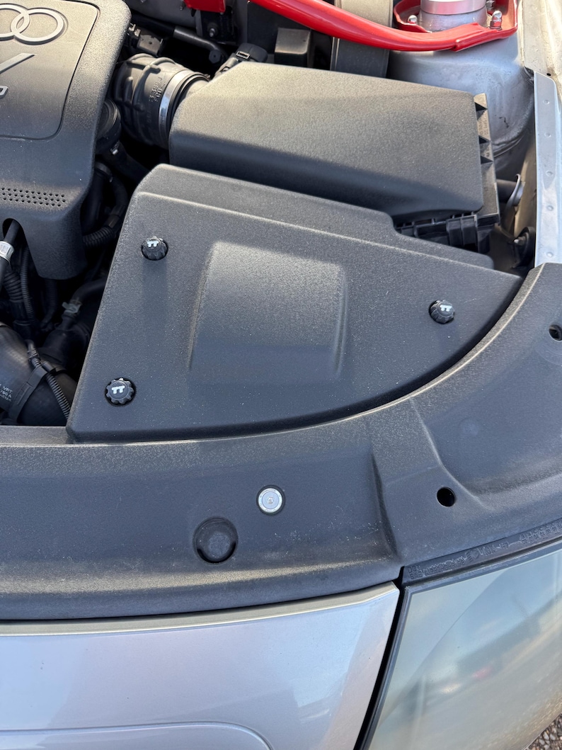 May include: A close-up view of a car engine compartment. The image shows a black plastic air intake box, silver metal components, and a red bar. The engine components are secured with silver bolts.