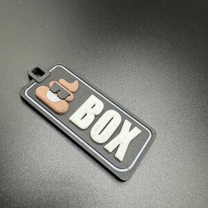 May include: Black and white luggage tag with the word "BOX" in white letters and a brown and black cartoon dog illustration.
