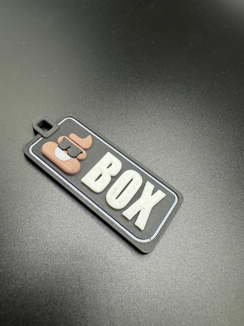 May include: Black and white luggage tag with the word "BOX" in white letters and a brown and black cartoon dog illustration.