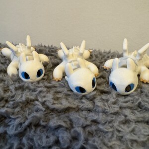 May include: Three white, 3D printed figurines of a dragon with blue eyes. The dragons are lying on their bellies with their wings spread out. They are on a gray, fuzzy surface.