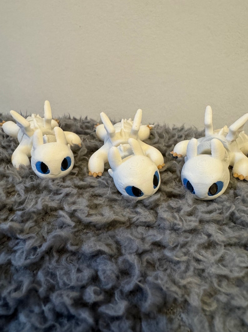 May include: Three white, 3D printed figurines of a dragon with blue eyes. The dragons are lying on their bellies with their wings spread out. They are on a gray, fuzzy surface.