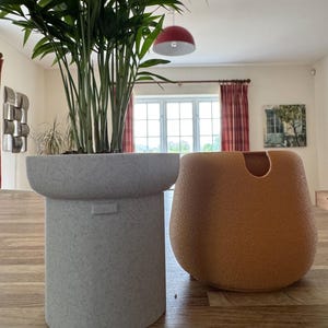 May include: Two decorative planters on a wooden surface. A tall, speckled gray planter holds a green plant. Beside it, an orange-brown, rounded planter with a curved opening. The room has a red pendant light and a window with red curtains.