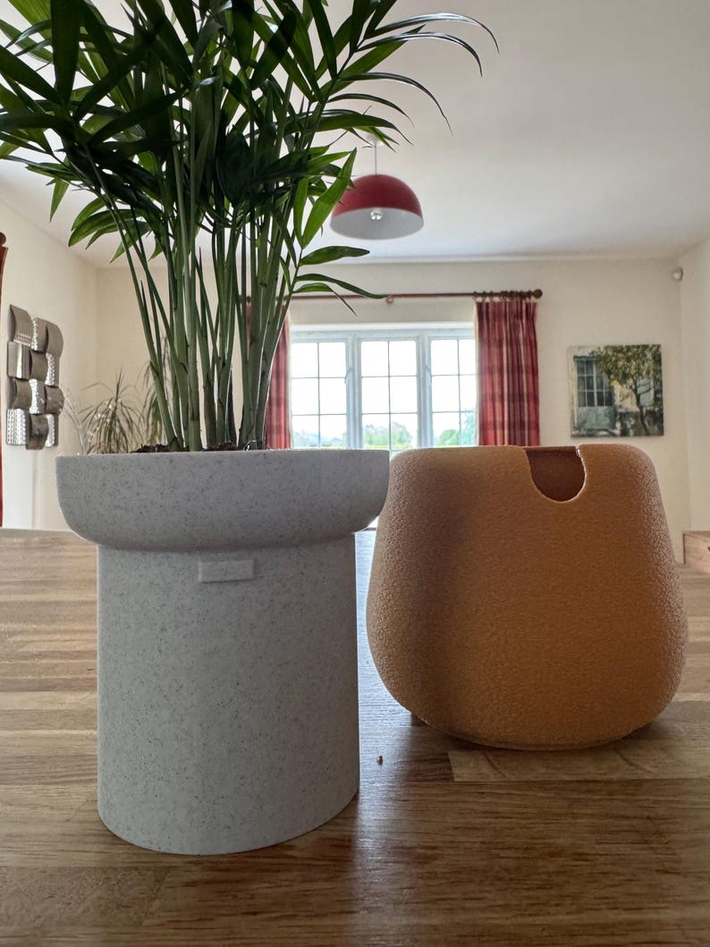 May include: Two decorative planters on a wooden surface. A tall, speckled gray planter holds a green plant. Beside it, an orange-brown, rounded planter with a curved opening. The room has a red pendant light and a window with red curtains.