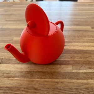 May include: A vibrant red teapot with a rounded body, a curved spout, and a small handle. The lid is slightly ajar, revealing the interior. This modern teapot is a stylish addition to any kitchen or tea set.