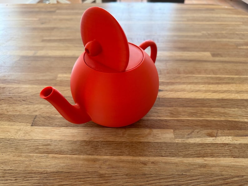 May include: A vibrant red teapot with a rounded body, a curved spout, and a small handle. The lid is slightly ajar, revealing the interior. This modern teapot is a stylish addition to any kitchen or tea set.