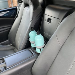 May include: Two light blue water bottles with black lids are in a car's cup holder. The car's interior features black leather seats with white stitching. The cup holder is gray. The car's interior is clean and well-maintained.