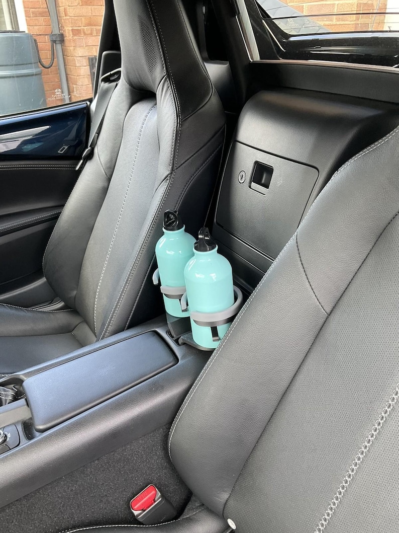 May include: Two light blue water bottles with black lids are in a car's cup holder. The car's interior features black leather seats with white stitching. The cup holder is gray. The car's interior is clean and well-maintained.