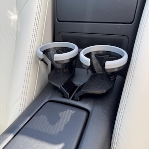 K&ouml;nnte beinhalten: Ein schwarzer Getr&auml;nkehalter f&uuml;r das Auto mit zwei kreisf&ouml;rmigen &Ouml;ffnungen, jeweils mit einem hellgrauen Rand. Der Getr&auml;nkehalter ist f&uuml;r den Einbau in die Mittelkonsole eines Autos konzipiert, wie auf dem Bild zu sehen.