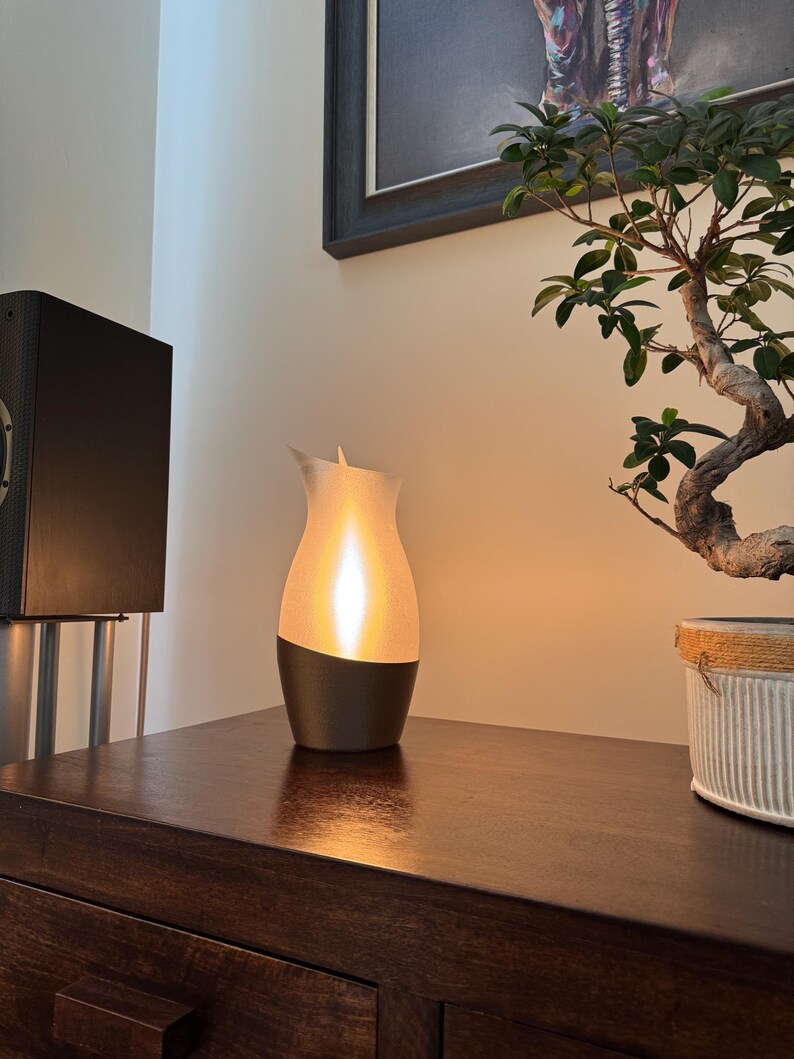 May include: A modern, decorative lamp with a warm, glowing light. The lamp has a dark base and a white, curved top. It sits on a dark wooden surface, next to a bonsai tree in a decorative pot. The lamp's design resembles a flame.