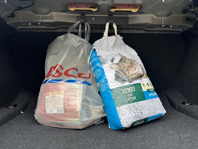 以下が含まれることがあります： 車のトランクにある2つの買い物袋。Tescoのロゴが入った透明なビニール袋には、包装された肉が入っています。もう一方の袋は、猫の画像と青と白のデザインのTesco猫用トイレ砂の袋です。