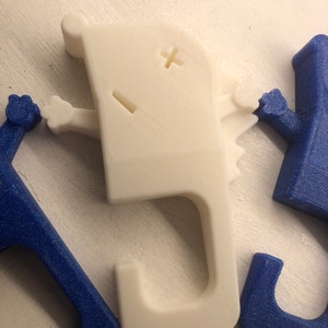 May include: White 3D printed hook with a cartoon face and arms. The hook is shaped like a letter J and has a small plus sign and a dash for eyes.