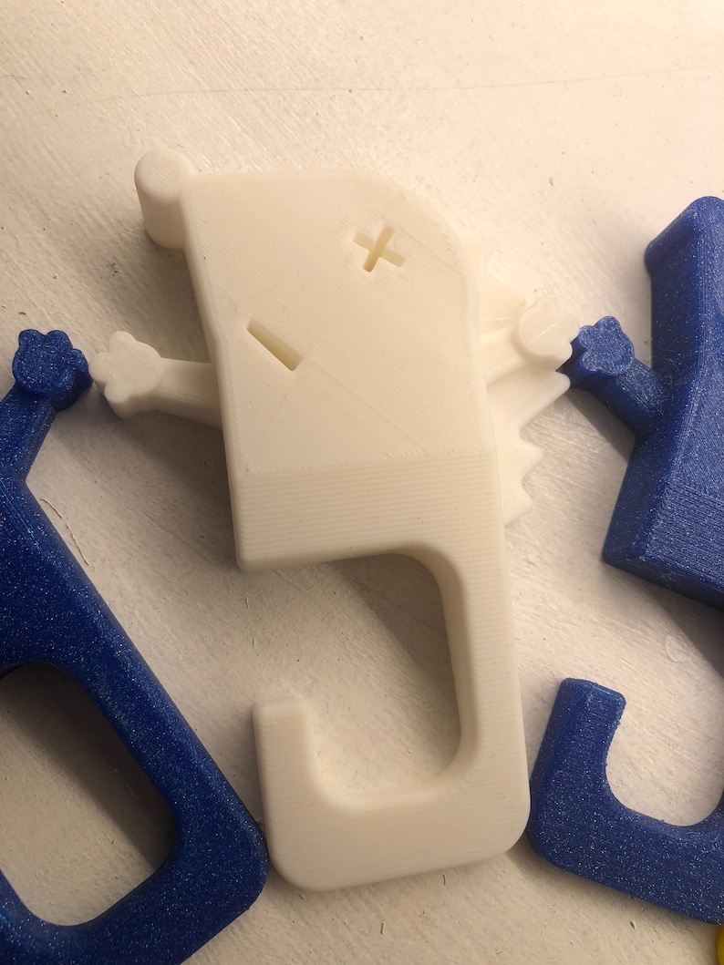 May include: White 3D printed hook with a cartoon face and arms. The hook is shaped like a letter J and has a small plus sign and a dash for eyes.