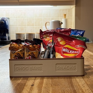 May include: A beige wooden snack organizer with compartments labeled "CHOC BARS" and "SNACKS." The organizer holds various packaged snacks, including chocolate bars and bags of chips, on a wooden countertop.