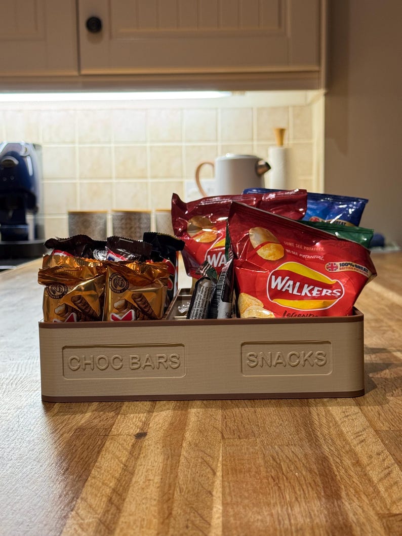 May include: A beige wooden snack organizer with compartments labeled "CHOC BARS" and "SNACKS." The organizer holds various packaged snacks, including chocolate bars and bags of chips, on a wooden countertop.