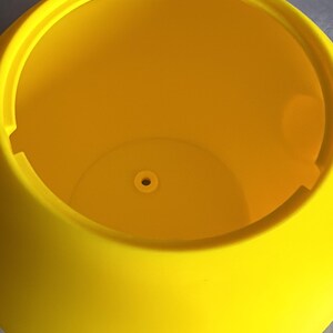 May include: A bright yellow, round plastic planter with a wide opening. The planter has a smooth, curved exterior and a small drainage hole at the bottom. The interior is a vibrant yellow color, and the planter is sitting on a gray surface.