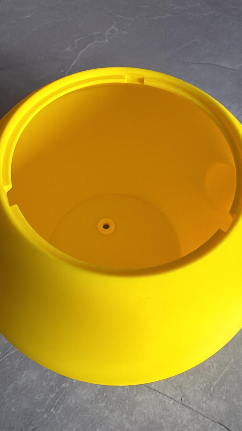 May include: A bright yellow, round plastic planter with a wide opening. The planter has a smooth, curved exterior and a small drainage hole at the bottom. The interior is a vibrant yellow color, and the planter is sitting on a gray surface.