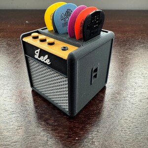 May include: A black and gray miniature guitar amplifier with a gold top and a label that reads "Lele". The amplifier has a slot on the top for holding guitar picks. There are five guitar picks in the slot, including a black pick, a red pick, a purple pick, a blue pick, and a yellow pick.