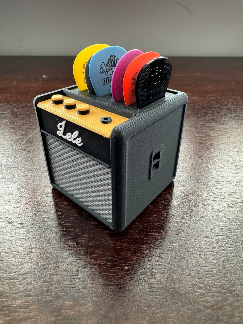 May include: A black and gray miniature guitar amplifier with a gold top and a label that reads "Lele". The amplifier has a slot on the top for holding guitar picks. There are five guitar picks in the slot, including a black pick, a red pick, a purple pick, a blue pick, and a yellow pick.