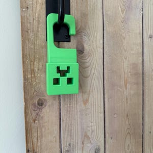 May include: A bright green door hanger featuring a pixelated design, reminiscent of a popular video game character. The hanger is attached to a black door handle, set against a wooden door with visible grain.