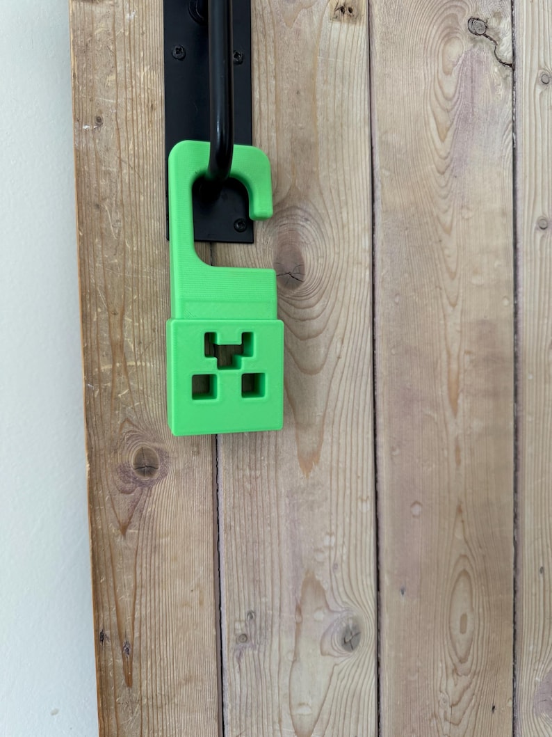 May include: A bright green door hanger featuring a pixelated design, reminiscent of a popular video game character. The hanger is attached to a black door handle, set against a wooden door with visible grain.