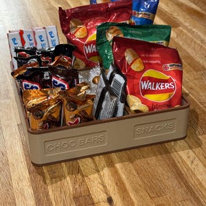 May include: A beige rectangular tray filled with various snacks. The tray is labeled "CHOC BARS" and "SNACKS". Inside are chocolate bars and bags of potato chips in red, green, and blue packaging. The chips are labeled "WALKERS".