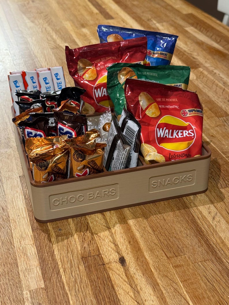 May include: A beige rectangular tray filled with various snacks. The tray is labeled "CHOC BARS" and "SNACKS". Inside are chocolate bars and bags of potato chips in red, green, and blue packaging. The chips are labeled "WALKERS".