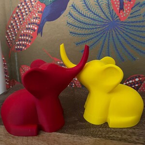 Op de afbeelding: Twee kleine olifantenfiguurtjes, een rode en een gele, staan ​​naast elkaar op een houten oppervlak. De rode olifant heeft zijn slurf omhoog en kijkt naar de gele olifant, die zijn slurf naar beneden heeft.