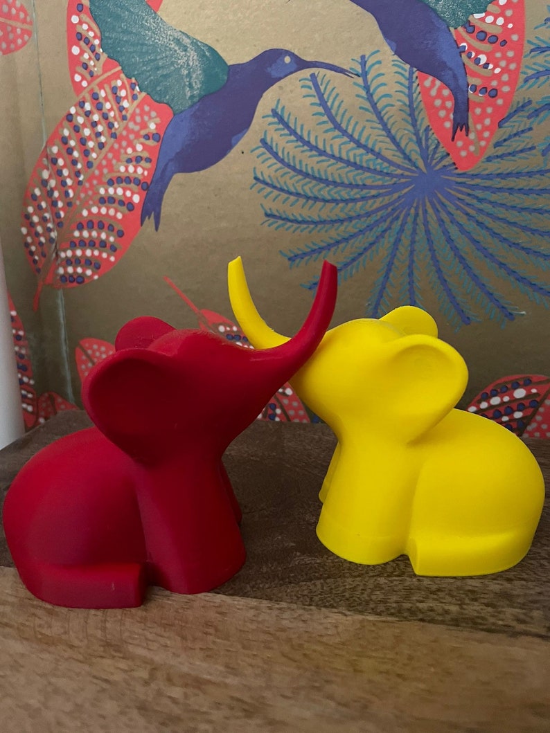 Op de afbeelding: Twee kleine olifantenfiguurtjes, een rode en een gele, staan ​​naast elkaar op een houten oppervlak. De rode olifant heeft zijn slurf omhoog en kijkt naar de gele olifant, die zijn slurf naar beneden heeft.