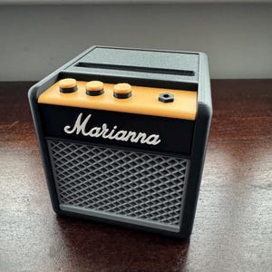May include: A black and orange miniature guitar amplifier with the word "Marianna" printed on the front. The amplifier has three knobs and a speaker grill.