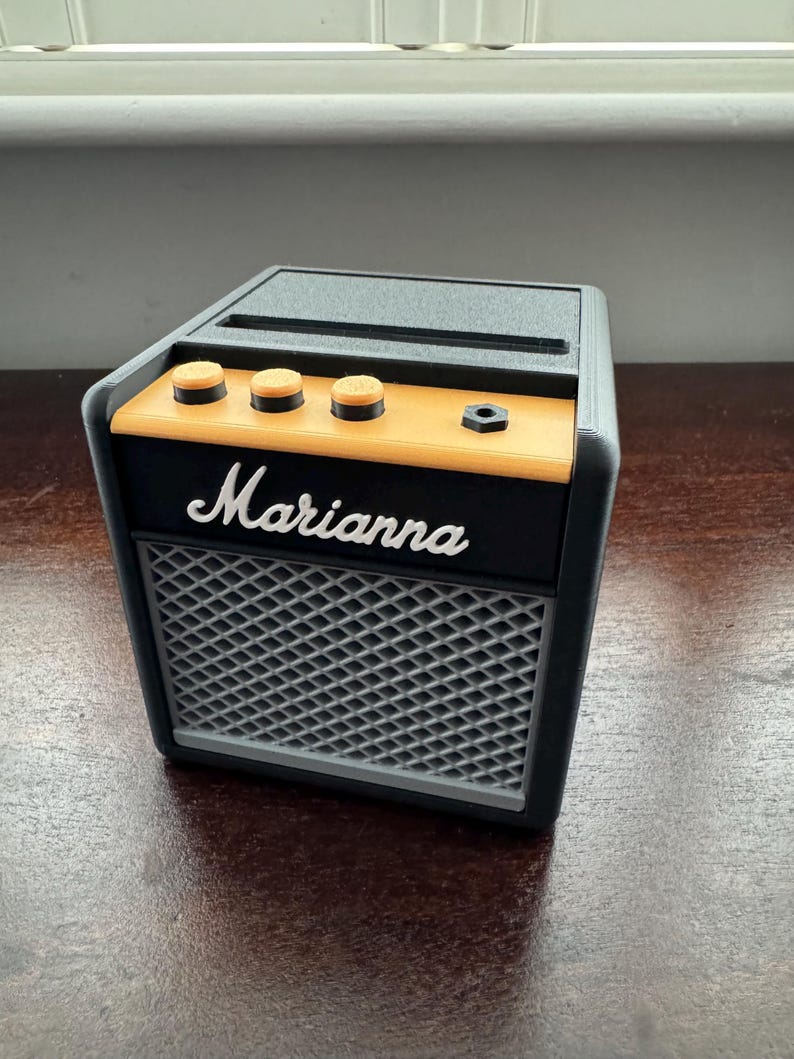 May include: A black and orange miniature guitar amplifier with the word "Marianna" printed on the front. The amplifier has three knobs and a speaker grill.