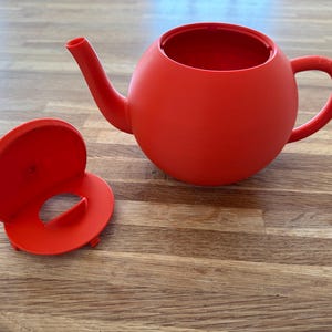 May include: A bright red teapot with a matching lid. The teapot has a rounded body, a curved spout, and a handle. The lid is circular with a central opening. The teapot and lid are made of a solid material.