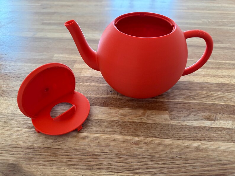 May include: A bright red teapot with a matching lid. The teapot has a rounded body, a curved spout, and a handle. The lid is circular with a central opening. The teapot and lid are made of a solid material.