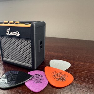 May include: A black Lewis guitar amplifier with a gray speaker grille and orange knobs. Four guitar picks are in front of the amplifier, including a black pick with the word "D'Addario" on it, a purple pick with the words "Tortex" and "U.S.A." on it, a white pick, and an orange pick with the word "Dunlop" on it.