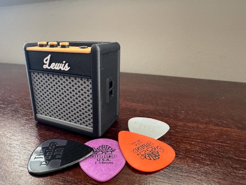 May include: A black Lewis guitar amplifier with a gray speaker grille and orange knobs. Four guitar picks are in front of the amplifier, including a black pick with the word "D'Addario" on it, a purple pick with the words "Tortex" and "U.S.A." on it, a white pick, and an orange pick with the word "Dunlop" on it.