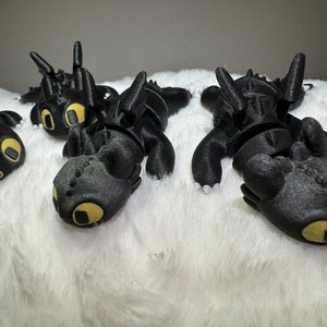 May include: Four black plush dragon toys with yellow eyes, lying on a white fluffy surface.